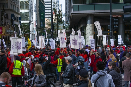 A large group of teachers gather on a city street in Chicago, holding protest signs and banners; some wear red shirts and yellow vests, while bystanders and media observe.
