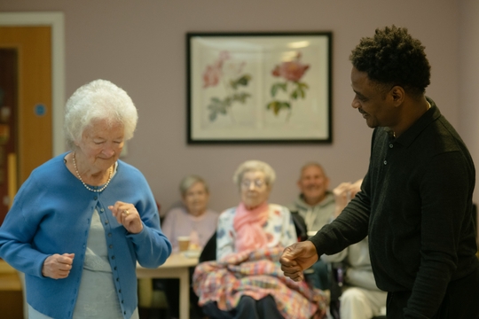 The image shows an elderly lady standing up and dancing, being watched by a row of elderly women sitting down. There is a younger man reaching out to her and smiling.