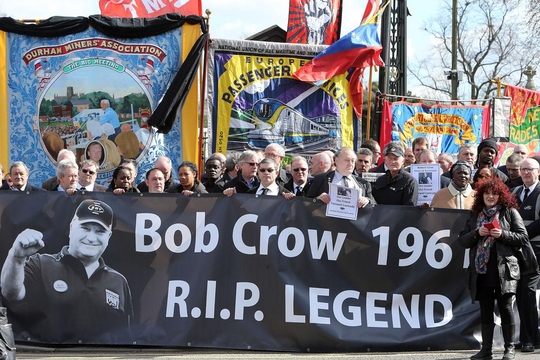 A group of people are participating in a memorial event, holding a banner honouring Bob Crow.